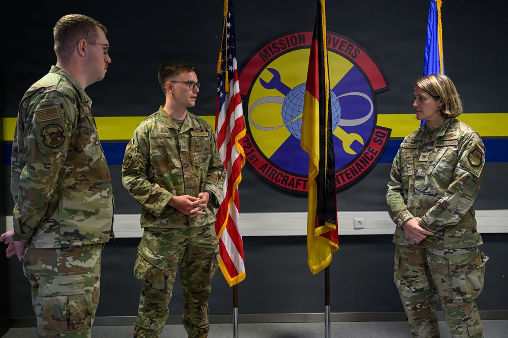 Three U.S. Air Force personnel in OCP (Operational Camouflage Pattern) uniforms are gathered in front of a unit emblem for the 321st Aircraft Maintenance Squadron, nicknamed 'Mission Movers.' The central figure appears to be speaking or being addressed in what looks like a formal military ceremony or recognition event. Flags including the U.S. flag and a unit/organizational guidon are displayed in the background.