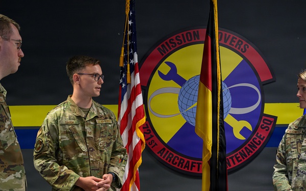 Three U.S. Air Force personnel in OCP (Operational Camouflage Pattern) uniforms are gathered in front of a unit emblem for the 321st Aircraft Maintenance Squadron, nicknamed 'Mission Movers.' The central figure appears to be speaking or being addressed in what looks like a formal military ceremony or recognition event. Flags including the U.S. flag and a unit/organizational guidon are displayed in the background.