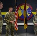 Three U.S. Air Force personnel in OCP (Operational Camouflage Pattern) uniforms are gathered in front of a unit emblem for the 321st Aircraft Maintenance Squadron, nicknamed 'Mission Movers.' The central figure appears to be speaking or being addressed in what looks like a formal military ceremony or recognition event. Flags including the U.S. flag and a unit/organizational guidon are displayed in the background.