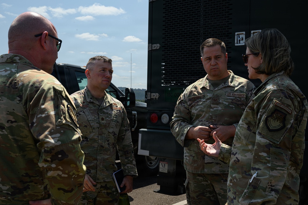 Four U.S. Air Force personnel in OCP (Operational Camouflage Pattern) uniforms are engaged in a discussion outdoors near military vehicles. Three individuals are facing one direction while one person gestures with their hand, suggesting a briefing or operational discussion. The setting appears to be a military installation or field environment.
