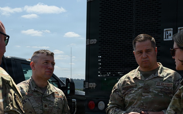 Four U.S. Air Force personnel in OCP (Operational Camouflage Pattern) uniforms are engaged in a discussion outdoors near military vehicles. Three individuals are facing one direction while one person gestures with their hand, suggesting a briefing or operational discussion. The setting appears to be a military installation or field environment.