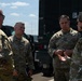 Four U.S. Air Force personnel in OCP (Operational Camouflage Pattern) uniforms are engaged in a discussion outdoors near military vehicles. Three individuals are facing one direction while one person gestures with their hand, suggesting a briefing or operational discussion. The setting appears to be a military installation or field environment.
