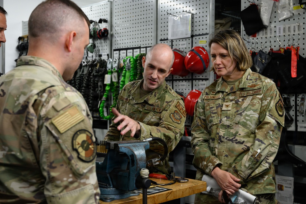 Three U.S. Air Force personnel in OCP (Operational Camouflage Pattern) uniforms are gathered around a workbench in what appears to be an equipment or maintenance shop. A male airman is gesturing toward a bench vise while explaining something to the other two personnel, one of whom is a female officer with a colonel's eagle insignia visible on her uniform. The background shows organized storage of safety and rigging equipment including harnesses, helmets, and straps.