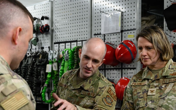 Three U.S. Air Force personnel in OCP (Operational Camouflage Pattern) uniforms are gathered around a workbench in what appears to be an equipment or maintenance shop. A male airman is gesturing toward a bench vise while explaining something to the other two personnel, one of whom is a female officer with a colonel's eagle insignia visible on her uniform. The background shows organized storage of safety and rigging equipment including harnesses, helmets, and straps.