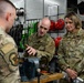 Three U.S. Air Force personnel in OCP (Operational Camouflage Pattern) uniforms are gathered around a workbench in what appears to be an equipment or maintenance shop. A male airman is gesturing toward a bench vise while explaining something to the other two personnel, one of whom is a female officer with a colonel's eagle insignia visible on her uniform. The background shows organized storage of safety and rigging equipment including harnesses, helmets, and straps.