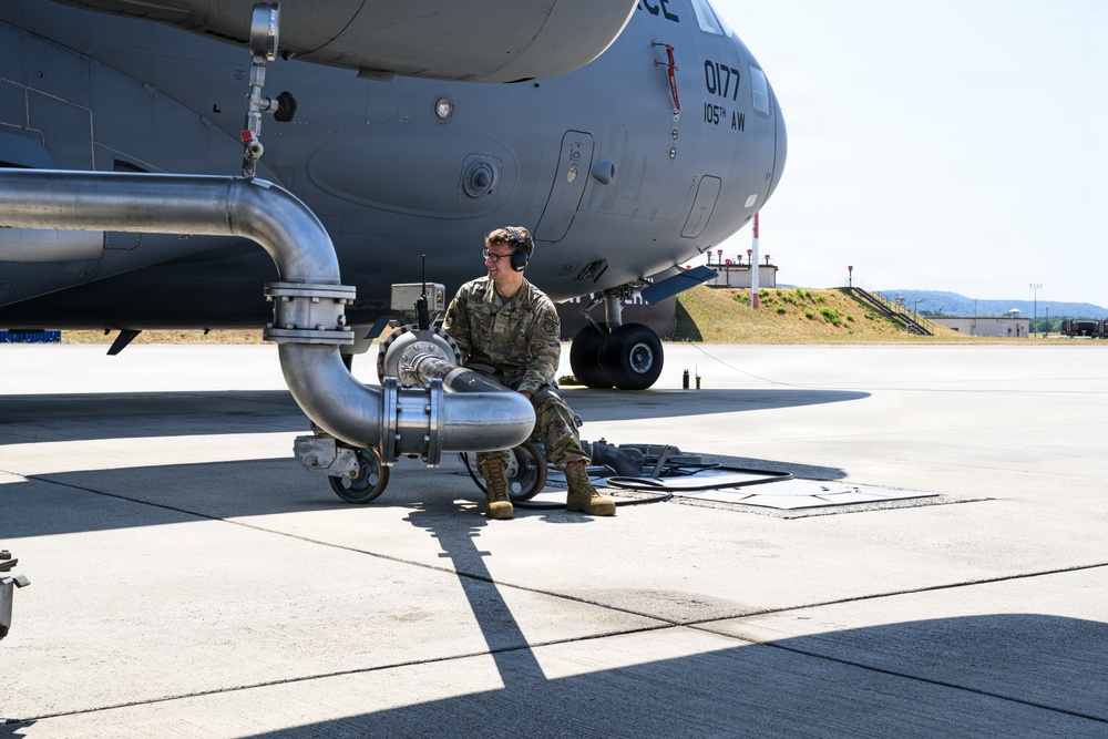 A military service member in OCP (Operational Camouflage Pattern) uniform is performing ground fueling operations on a large military transport aircraft. The aircraft is marked '0177' and '105th AW' (Airlift Wing), identifiable as a C-17 Globemaster III. The service member is crouched near large fuel pipe assemblies connected to an underground fuel pit on a concrete flight line.