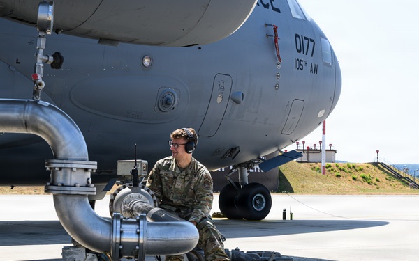 A military service member in OCP (Operational Camouflage Pattern) uniform is performing ground fueling operations on a large military transport aircraft. The aircraft is marked '0177' and '105th AW' (Airlift Wing), identifiable as a C-17 Globemaster III. The service member is crouched near large fuel pipe assemblies connected to an underground fuel pit on a concrete flight line.