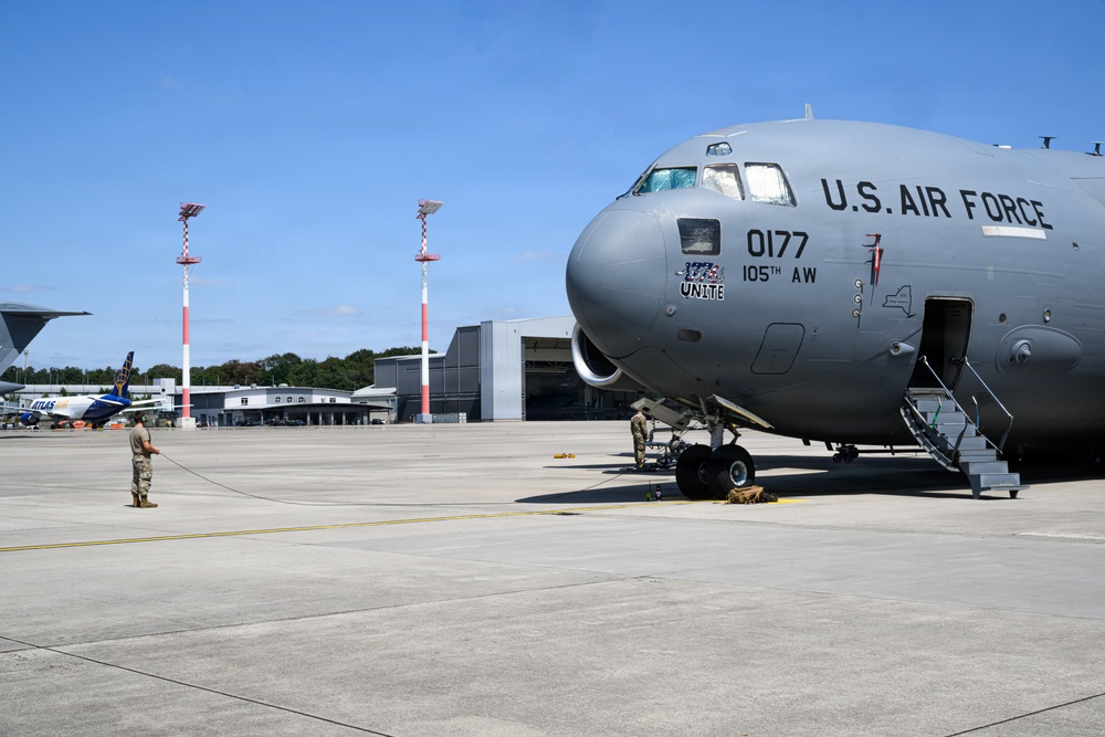 A U.S. Air Force C-17 Globemaster III military transport aircraft, tail number 0177, assigned to the 105th Airlift Wing, is parked on an airport tarmac. Ground crew personnel are visible performing pre/post-flight servicing activities around the aircraft. The scene appears to be at a military or joint-use airfield, with hangars and other aircraft visible in the background.