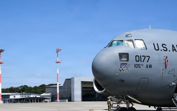 A U.S. Air Force C-17 Globemaster III military transport aircraft, tail number 0177, assigned to the 105th Airlift Wing, is parked on an airport tarmac. Ground crew personnel are visible performing pre/post-flight servicing activities around the aircraft. The scene appears to be at a military or joint-use airfield, with hangars and other aircraft visible in the background.