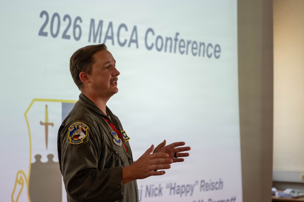 A military officer in a flight suit is presenting at the 2026 MACA Conference. The speaker is identified by a name banner as Maj Nick 'Happy' Reisch, and he is gesturing with his hands while speaking in front of a projected slide. He wears patches consistent with a U.S. Air Force fighter wing unit.