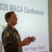 A military officer in a flight suit is presenting at the 2026 MACA Conference. The speaker is identified by a name banner as Maj Nick 'Happy' Reisch, and he is gesturing with his hands while speaking in front of a projected slide. He wears patches consistent with a U.S. Air Force fighter wing unit.