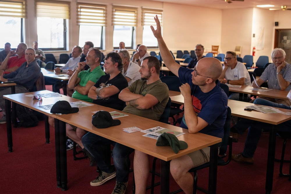 A group of predominantly male adults are seated in a classroom or meeting room setting, attending what appears to be a seminar, briefing, or training session. One bald man wearing glasses and a dark blue t-shirt has his hand raised, likely asking a question or responding to the presenter. The room contains multiple rows of desks with printed materials distributed among attendees.