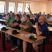 A group of predominantly male adults are seated in a classroom or meeting room setting, attending what appears to be a seminar, briefing, or training session. One bald man wearing glasses and a dark blue t-shirt has his hand raised, likely asking a question or responding to the presenter. The room contains multiple rows of desks with printed materials distributed among attendees.