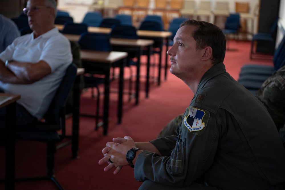 A military personnel member wearing an olive green flight suit with a unit patch is seated in what appears to be a briefing room or classroom, attentively listening. The room contains multiple empty blue chairs and desks arranged in rows. A civilian in a white shirt is visible in the background with arms crossed.