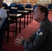 A military personnel member wearing an olive green flight suit with a unit patch is seated in what appears to be a briefing room or classroom, attentively listening. The room contains multiple empty blue chairs and desks arranged in rows. A civilian in a white shirt is visible in the background with arms crossed.