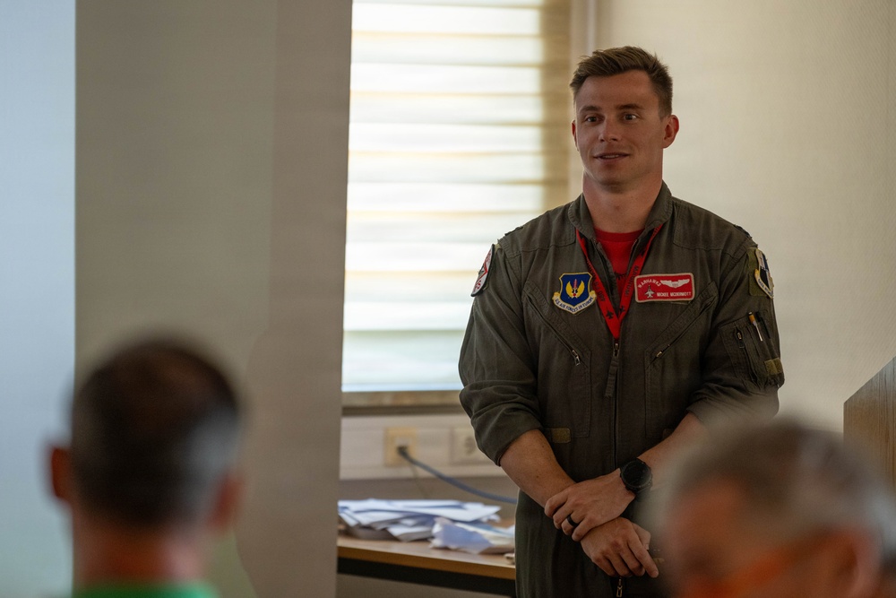 A young male U.S. Air Force aviator in an olive green flight suit stands in what appears to be a briefing room or classroom setting, addressing an audience. He is wearing a name tag, wing insignia, and unit patches on his flight suit, with a red shirt visible underneath. The setting suggests a formal or semi-formal military briefing or presentation.