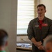 A young male U.S. Air Force aviator in an olive green flight suit stands in what appears to be a briefing room or classroom setting, addressing an audience. He is wearing a name tag, wing insignia, and unit patches on his flight suit, with a red shirt visible underneath. The setting suggests a formal or semi-formal military briefing or presentation.