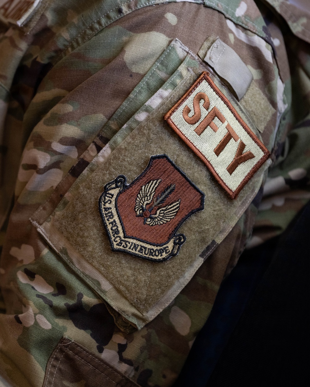 Close-up photograph of a U.S. military uniform sleeve displaying two patches on a MultiCam/OCP camouflage uniform. The patches identify the wearer as affiliated with U.S. Air Forces in Europe (USAFE) and carrying a 'SFTY' (Safety) functional designation. The image is a detail shot focused on the left shoulder area of the uniform.