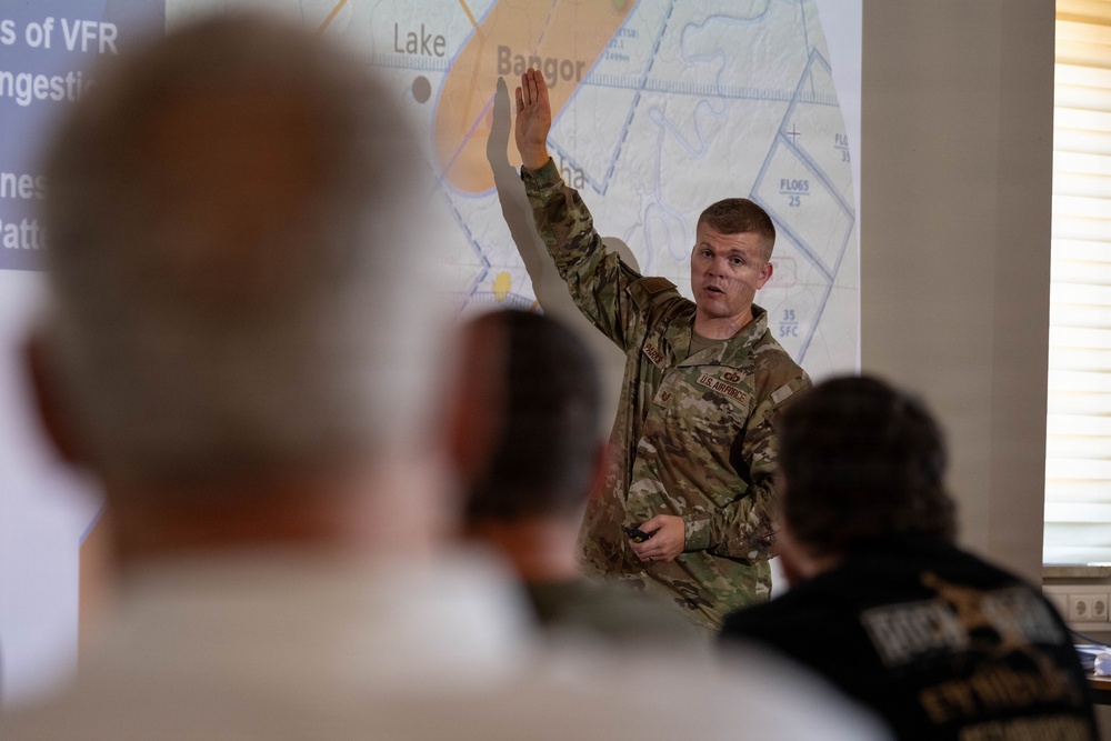 A U.S. Air Force servicemember in OCP (Operational Camouflage Pattern) uniform is delivering a briefing or presentation to a seated audience. The presenter has one hand raised and is holding a remote clicker in the other hand, gesturing toward a projected map showing the Bangor, Maine area with airspace designations. The slide content references VFR (Visual Flight Rules) topics including traffic patterns.