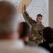 A U.S. Air Force servicemember in OCP (Operational Camouflage Pattern) uniform is delivering a briefing or presentation to a seated audience. The presenter has one hand raised and is holding a remote clicker in the other hand, gesturing toward a projected map showing the Bangor, Maine area with airspace designations. The slide content references VFR (Visual Flight Rules) topics including traffic patterns.
