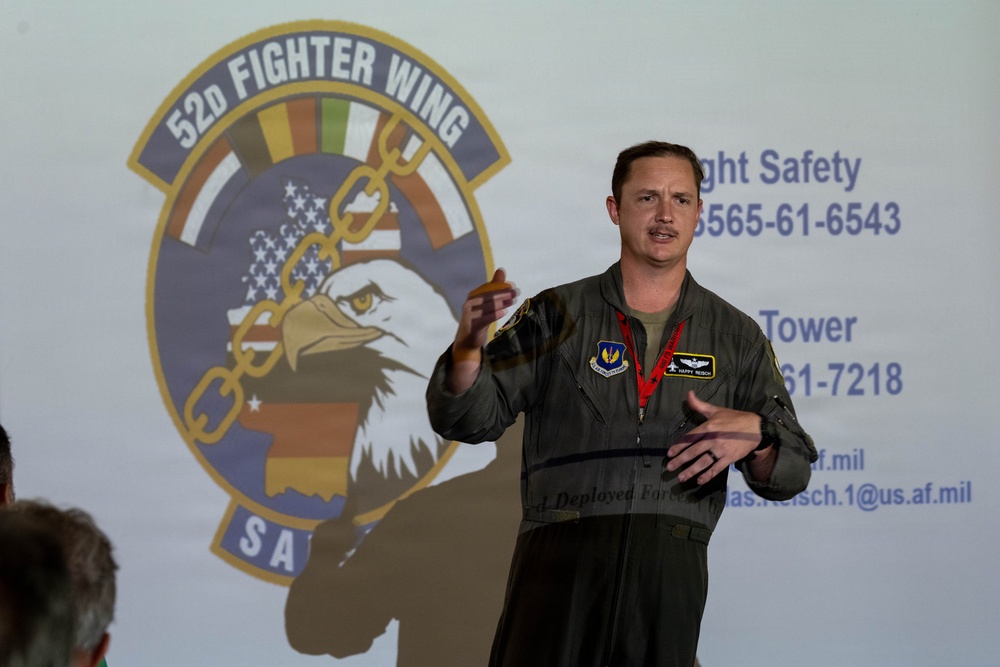 A U.S. Air Force officer in a flight suit is presenting or briefing in front of a projected screen displaying the 52d Fighter Wing emblem and contact information. The speaker appears to be addressing an audience, with at least one other person partially visible in the foreground. The setting appears to be a hangar or briefing room at a military installation.