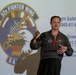 A U.S. Air Force officer in a flight suit is presenting or briefing in front of a projected screen displaying the 52d Fighter Wing emblem and contact information. The speaker appears to be addressing an audience, with at least one other person partially visible in the foreground. The setting appears to be a hangar or briefing room at a military installation.