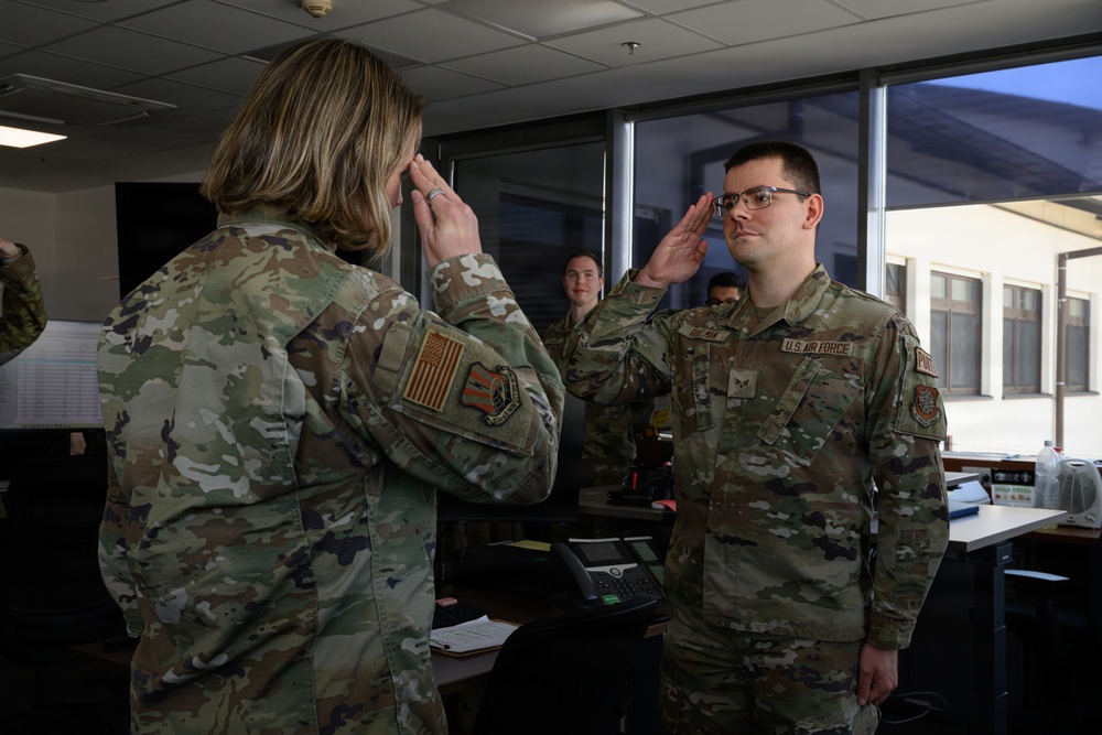 Two U.S. Air Force personnel in OCP (Operational Camouflage Pattern) uniforms are exchanging salutes in what appears to be an indoor office or operations center environment. The individual facing the camera wears a 'U.S. Air Force' tape and rank insignia visible on their uniform. Additional personnel are visible in the background observing the exchange.
