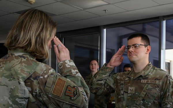 Two U.S. Air Force personnel in OCP (Operational Camouflage Pattern) uniforms are exchanging salutes in what appears to be an indoor office or operations center environment. The individual facing the camera wears a 'U.S. Air Force' tape and rank insignia visible on their uniform. Additional personnel are visible in the background observing the exchange.