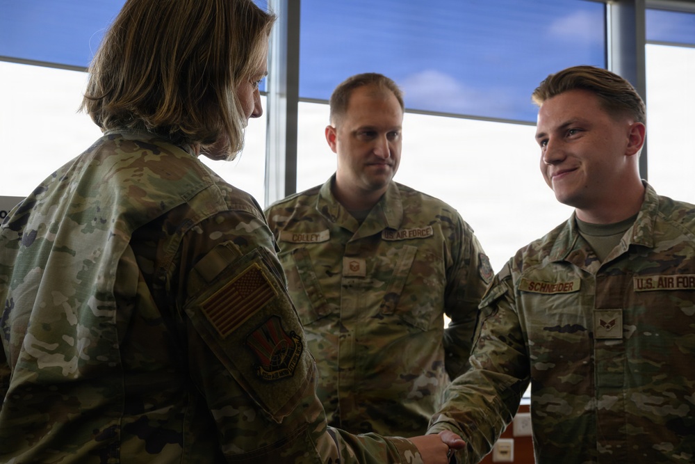 Three U.S. Air Force personnel in OCP (Operational Camouflage Pattern) uniforms are engaged in what appears to be a formal handshake interaction, likely a recognition or greeting ceremony. A senior officer (facing away) is shaking hands with Airman Schneider on the right, while a service member named Colley observes from the center. The setting appears to be an indoor facility with large windows in the background.