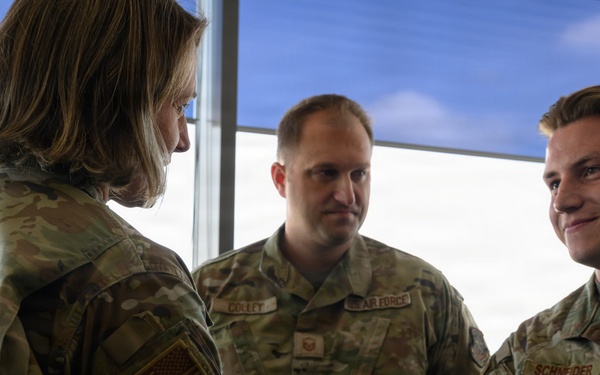 Three U.S. Air Force personnel in OCP (Operational Camouflage Pattern) uniforms are engaged in what appears to be a formal handshake interaction, likely a recognition or greeting ceremony. A senior officer (facing away) is shaking hands with Airman Schneider on the right, while a service member named Colley observes from the center. The setting appears to be an indoor facility with large windows in the background.
