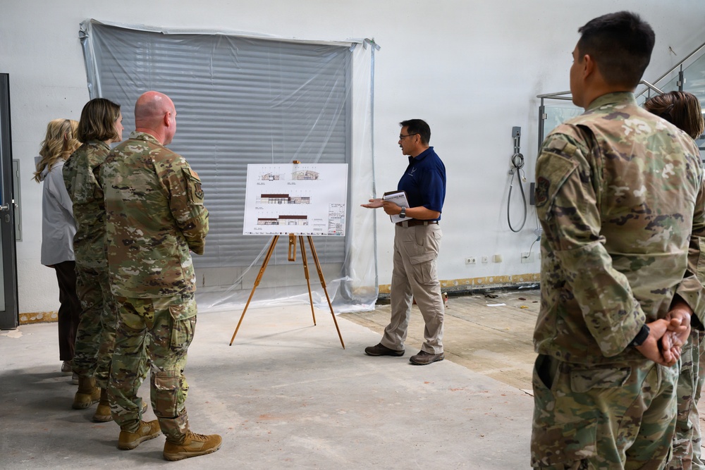 A civilian presenter in a blue polo shirt is briefing a group of military personnel in camouflage uniforms inside what appears to be a building under renovation or construction. The presenter is standing next to an easel displaying architectural or engineering drawings, likely floor plans or building elevations. The setting suggests a construction site walkthrough or project review meeting.