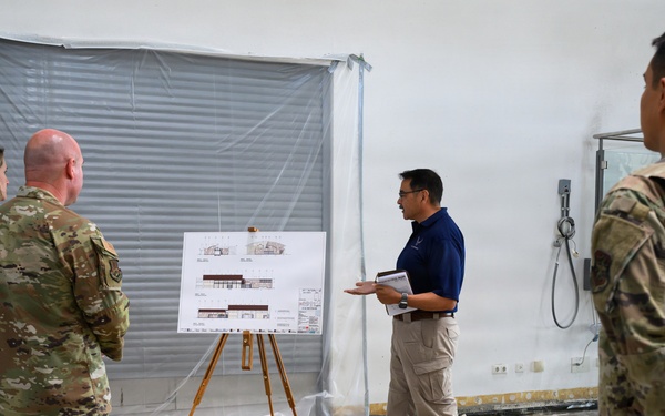 A civilian presenter in a blue polo shirt is briefing a group of military personnel in camouflage uniforms inside what appears to be a building under renovation or construction. The presenter is standing next to an easel displaying architectural or engineering drawings, likely floor plans or building elevations. The setting suggests a construction site walkthrough or project review meeting.