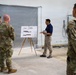 A civilian presenter in a blue polo shirt is briefing a group of military personnel in camouflage uniforms inside what appears to be a building under renovation or construction. The presenter is standing next to an easel displaying architectural or engineering drawings, likely floor plans or building elevations. The setting suggests a construction site walkthrough or project review meeting.