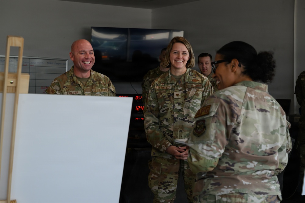 Three U.S. Air Force personnel in OCP (Operational Camouflage Pattern) uniforms are engaged in a conversation in what appears to be a military operations or briefing room. Two individuals facing the camera — name tapes reading 'FINCHER' and 'POLSINELLI' — are smiling and laughing. A whiteboard on an easel is visible in the foreground, and digital clocks and a large display screen are visible in the background.