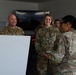 Three U.S. Air Force personnel in OCP (Operational Camouflage Pattern) uniforms are engaged in a conversation in what appears to be a military operations or briefing room. Two individuals facing the camera — name tapes reading 'FINCHER' and 'POLSINELLI' — are smiling and laughing. A whiteboard on an easel is visible in the foreground, and digital clocks and a large display screen are visible in the background.