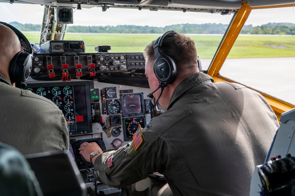 Two military aviators are seated in the cockpit of a large military aircraft, likely a tanker or transport such as a KC-135 or C-17, on an airfield tarmac. Both crew members are wearing olive drab flight suits and aviation headsets, with one identified as wearing Bose headphones and displaying a US flag patch on their sleeve. The cockpit features a mix of analog gauges and modern digital displays, suggesting an upgraded avionics suite.