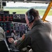 Two military aviators are seated in the cockpit of a large military aircraft, likely a tanker or transport such as a KC-135 or C-17, on an airfield tarmac. Both crew members are wearing olive drab flight suits and aviation headsets, with one identified as wearing Bose headphones and displaying a US flag patch on their sleeve. The cockpit features a mix of analog gauges and modern digital displays, suggesting an upgraded avionics suite.