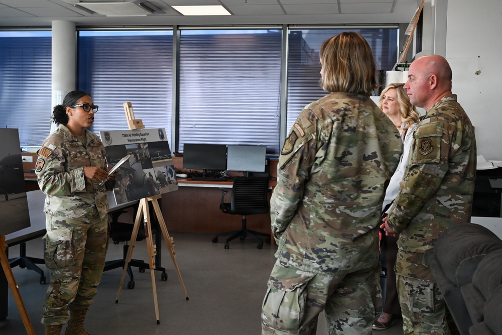 A military briefing is taking place in an office setting where a female service member in OCP (Operational Camouflage Pattern) uniform is presenting information using a display board on an easel. Three individuals — two in military uniforms and one civilian woman — are observing the presentation. The display board appears to reference the '728th Air Mobility Squadron Maintenance Flight.'