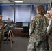 A military briefing is taking place in an office setting where a female service member in OCP (Operational Camouflage Pattern) uniform is presenting information using a display board on an easel. Three individuals — two in military uniforms and one civilian woman — are observing the presentation. The display board appears to reference the '728th Air Mobility Squadron Maintenance Flight.'