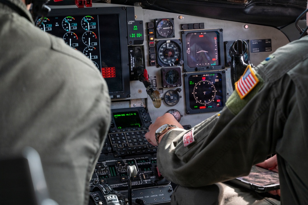 Two military aviators are seated in the cockpit of a large military aircraft, likely a transport or tanker aircraft such as a KC-135 or C-17. The right-seat pilot is actively operating the center console controls, wearing a flight suit with a US flag patch visible on the right shoulder. The instrument panel displays a mix of analog gauges and modern digital multifunction displays.