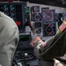 Two military aviators are seated in the cockpit of a large military aircraft, likely a transport or tanker aircraft such as a KC-135 or C-17. The right-seat pilot is actively operating the center console controls, wearing a flight suit with a US flag patch visible on the right shoulder. The instrument panel displays a mix of analog gauges and modern digital multifunction displays.