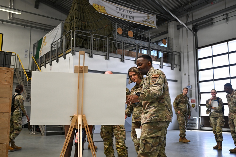 Military personnel in camouflage uniforms are gathered inside a large warehouse or hangar facility. Two service members in the foreground are examining a blank white board mounted on a wooden easel. A banner in the background reads '20 AMS Ramp 7' (reversed), suggesting this is a U.S. Air Force Air Mobility Squadron facility.