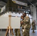 Military personnel in camouflage uniforms are gathered inside a large warehouse or hangar facility. Two service members in the foreground are examining a blank white board mounted on a wooden easel. A banner in the background reads '20 AMS Ramp 7' (reversed), suggesting this is a U.S. Air Force Air Mobility Squadron facility.