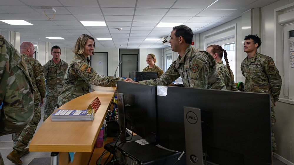 A group of military personnel in OCP (Operational Camouflage Pattern) uniforms are gathered in what appears to be an administrative or operations office. A female service member is shaking hands with a male service member across a reception-style desk equipped with Dell computer monitors. The setting suggests an official visit, briefing, or unit inspection.