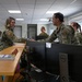 A group of military personnel in OCP (Operational Camouflage Pattern) uniforms are gathered in what appears to be an administrative or operations office. A female service member is shaking hands with a male service member across a reception-style desk equipped with Dell computer monitors. The setting suggests an official visit, briefing, or unit inspection.