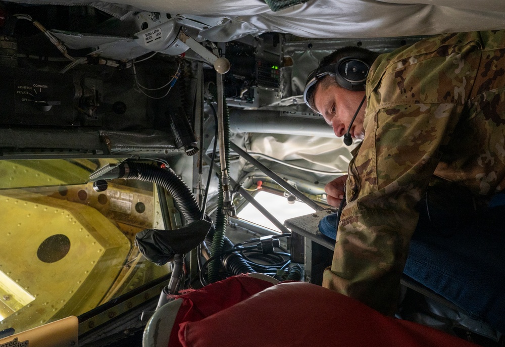 A military personnel member wearing multicam/OCP camouflage uniform and a communications headset is working inside what appears to be a military aircraft, likely performing maintenance or inspection duties. The individual is leaning into a confined interior space surrounded by wiring, structural components, and mechanical systems. The setting appears to be the interior boom operator or maintenance access area of a large military aircraft.