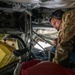 A military personnel member wearing multicam/OCP camouflage uniform and a communications headset is working inside what appears to be a military aircraft, likely performing maintenance or inspection duties. The individual is leaning into a confined interior space surrounded by wiring, structural components, and mechanical systems. The setting appears to be the interior boom operator or maintenance access area of a large military aircraft.