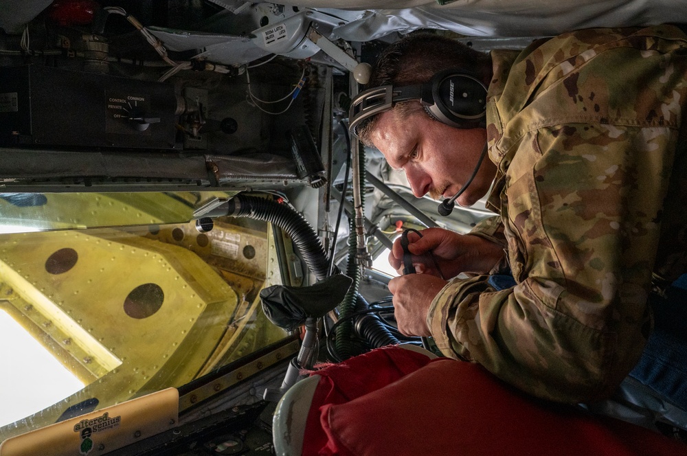 A military personnel member in multicam/OCP camouflage uniform is performing maintenance or technical work inside what appears to be a military aircraft. The individual is wearing a Bose headset with a boom microphone and is closely inspecting or manipulating cables and connectors in a confined compartment. The workspace contains various aircraft components including wiring harnesses, structural panels, and control equipment.