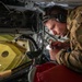 A military personnel member in multicam/OCP camouflage uniform is performing maintenance or technical work inside what appears to be a military aircraft. The individual is wearing a Bose headset with a boom microphone and is closely inspecting or manipulating cables and connectors in a confined compartment. The workspace contains various aircraft components including wiring harnesses, structural panels, and control equipment.