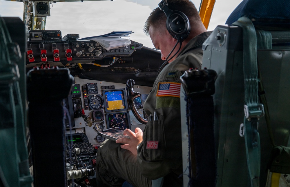 A U.S. military aviator is seated in the cockpit of a large military aircraft, reviewing a tablet device while in flight. The pilot is wearing a flight suit with a U.S. flag patch, Bose aviation headset, and has access to an extensive array of analog and digital cockpit instruments. The cockpit configuration and instrument panel suggest this is likely a large strategic aircraft such as a B-52 or KC-135.